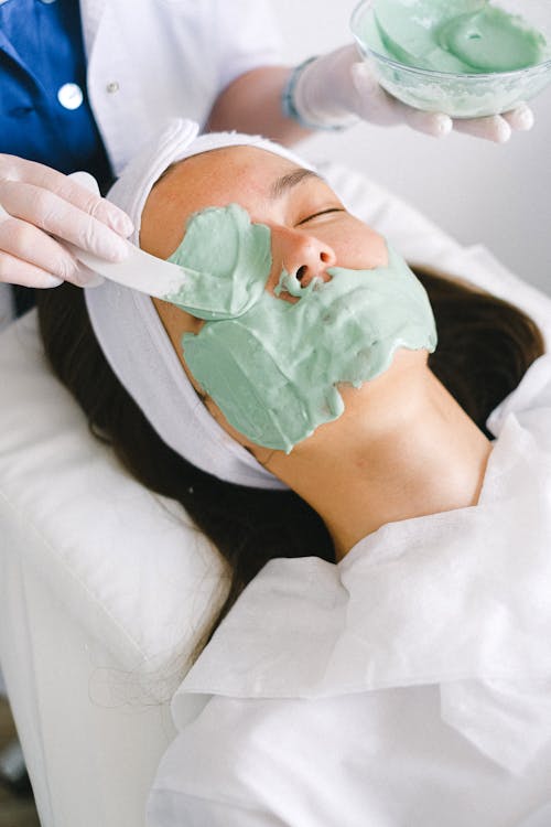 High angle professional female beautician applying moisturizing facial mask on serene female customer face skin in light contemporary beauty clinic