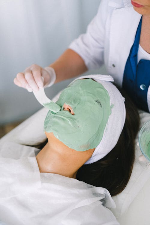 From above anonymous female beauty specialist applying revitalizing facial mask on female client face skin in light modern spa clinic