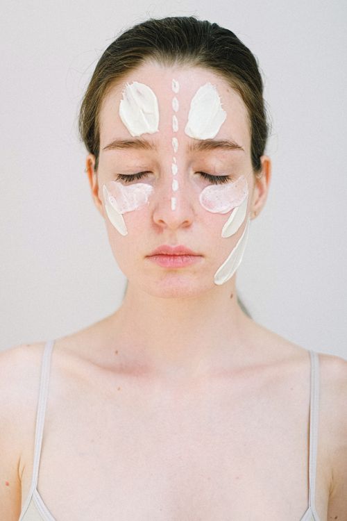 Free Young female model with perfect skin applying cosmetic cream on face while standing with closed eyes on white background Stock Photo