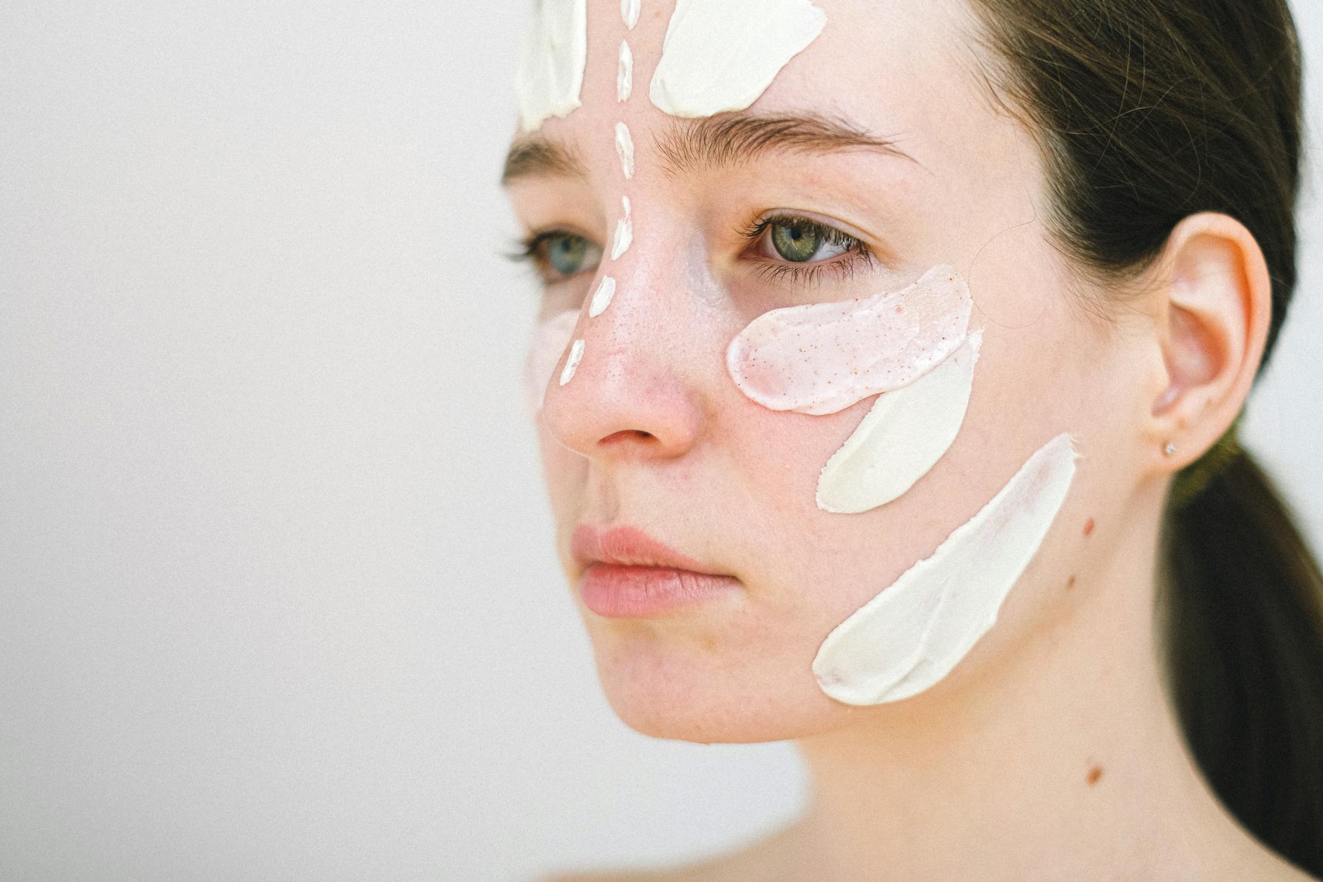 Side view of crop female model spreading white cream on face during skincare procedure on white background