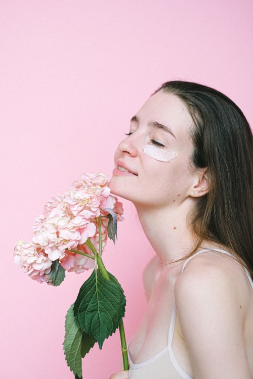 Free Side view of smiling young woman smelling fresh pink hydrangea flower while standing with cream under closed eyes on pink background Stock Photo