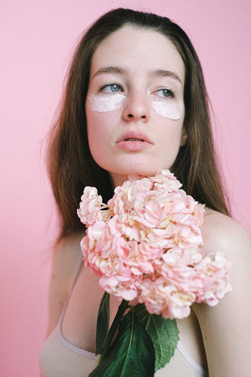 Thoughtful young female model with facial cream under eyes holding colorful flower