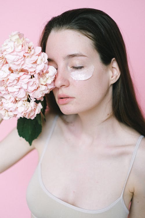 Young brunette in casual top standing on pink background with cosmetic mask on soft skin and hiding face behind pink hydrangea