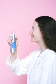 一位深色头发、身穿白色长袍的积极女性侧面图，粉红色背景中，脸上正喷洒着保湿化妆品
