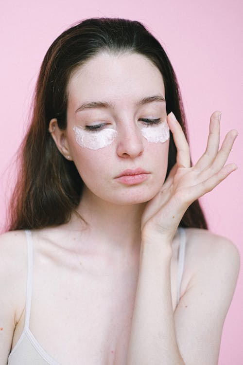 Free Young woman with dark hair in top applying moisturizing facial cream under eyes against pink background in studio Stock Photo