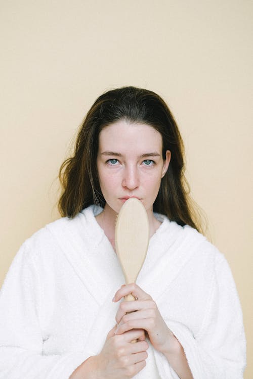 Calm young female with dark hair in white robe standing with wooden brush for anti cellulite massage in studio and looking at camera against beige background
