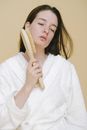 Calm female model with brown hair in white robe standing with closed eyes and touching face with brush for anti aging massage against beige background