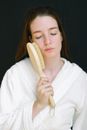 Young female with brown hair in white bathrobe standing with closed eyes and touching face with wooden brush for dry massage against black background