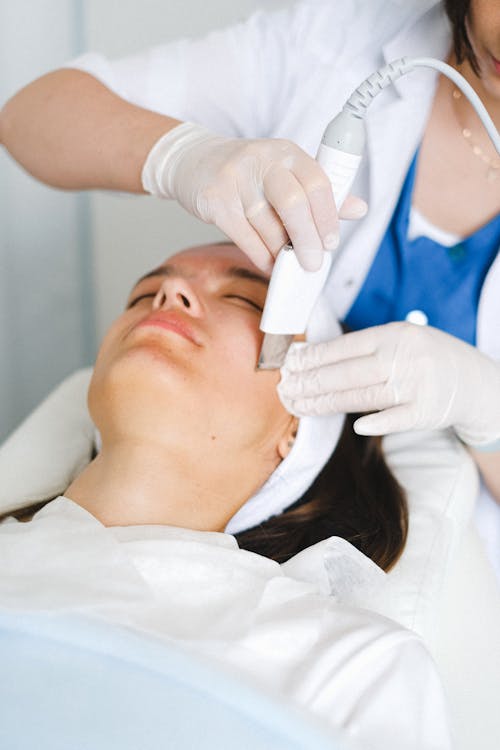 Crop unrecognizable female cosmetologist in medical uniform and gloves doing ultrasonic face cleaning procedure in beauty clinic