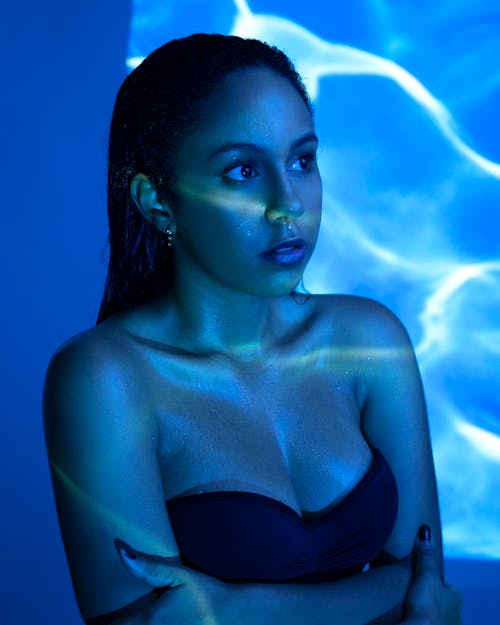 Young female with dark hair with naked shoulders standing with folded hands under blue neon illumination while looking away pensively in studio inside
