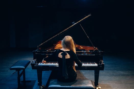 Woman playing a grand piano on stage, captured from behind, in a dimly lit performance setting.