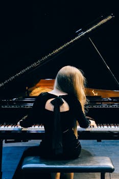 A woman in a black dress elegantly plays a grand piano on stage, showcasing musical talent.