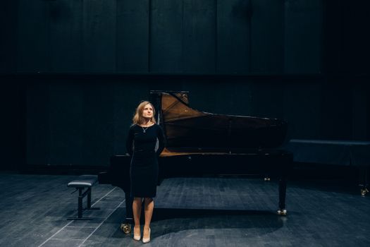 Woman in black dress standing by a grand piano in a concert hall.
