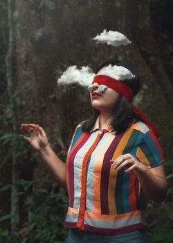 一张超现实照片中，一位蒙着眼睛的女人漂浮在森林里，周围环绕着云朵