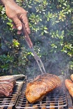 Hand grilling juicy meats on charcoal grill outdoors, creating a smokey flavor.