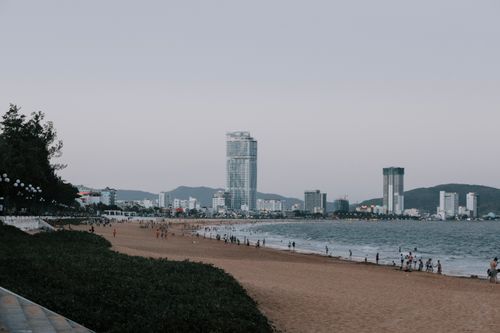 Free Distant silhouettes of people walking on sandy beach near rippling azure ocean against modern buildings on cloudless day in summer Stock Photo