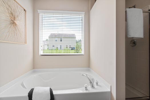 Modern bathroom featuring a white bathtub, window with blinds, and decorative wall art.