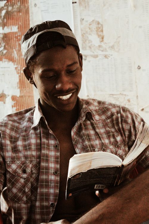 Free Cheerful African American male in hat in checkered cloth smiling and reading book while sitting near shabby wall in light room Stock Photo