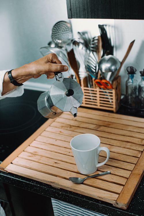Free From above of anonymous person pouring hot coffee from metal teapot into cup placed on wooden tray near utensils in kitchen Stock Photo