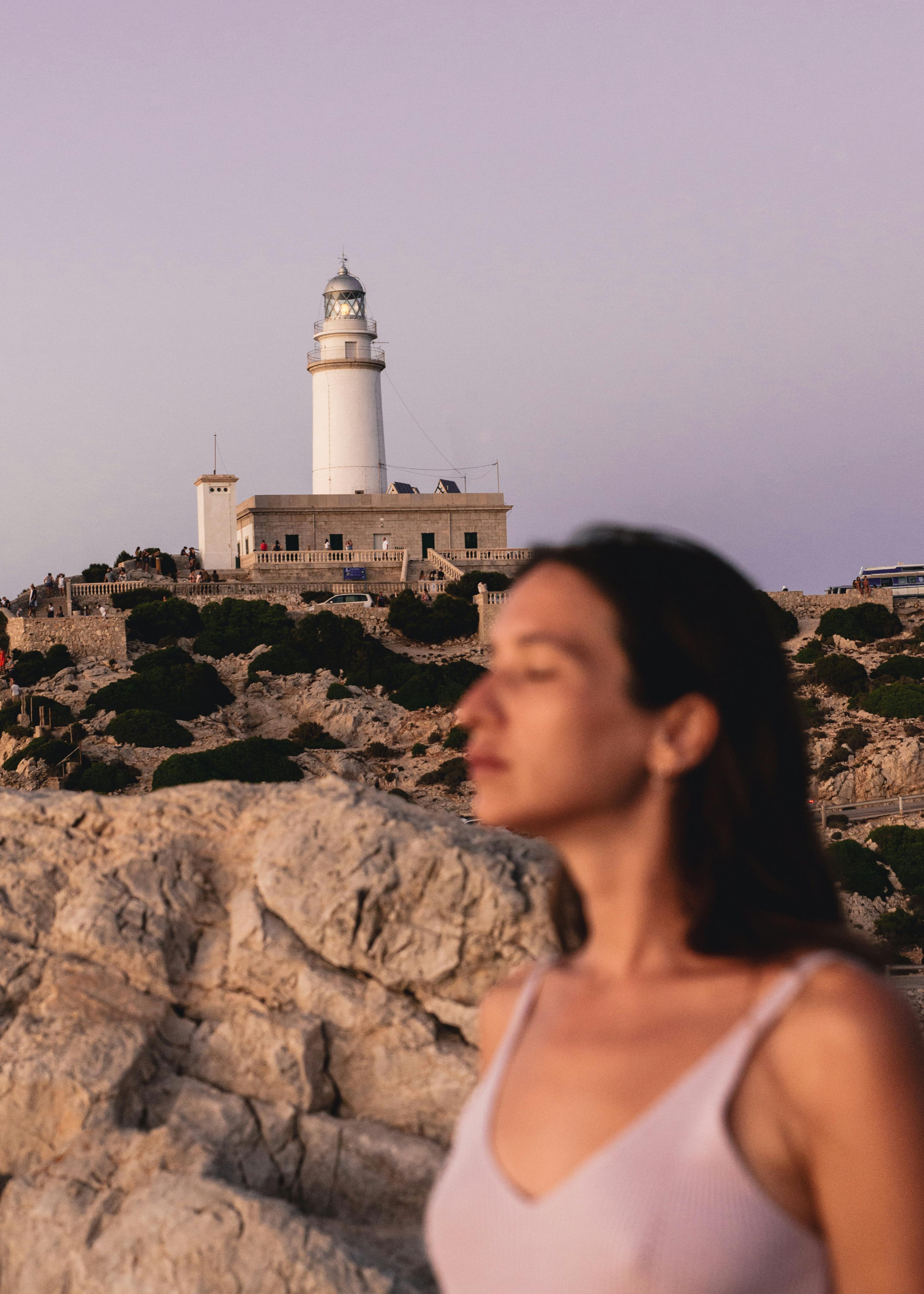A woman stands in front of the Alcanada Lighthouse at sunset in Pollença, Spain.