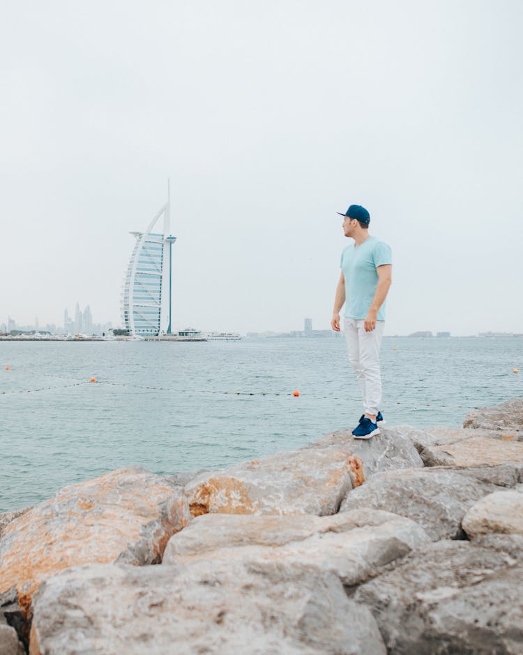 A Man On A Rocky Shore With A View Of The Burj Al Arab In Dubai