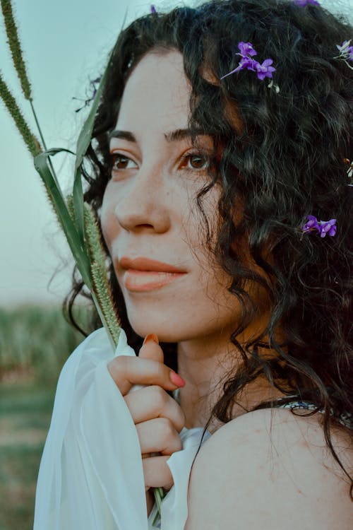 Dreamy woman with flower petal in wavy dark hair touching grass and looking away in daytime
