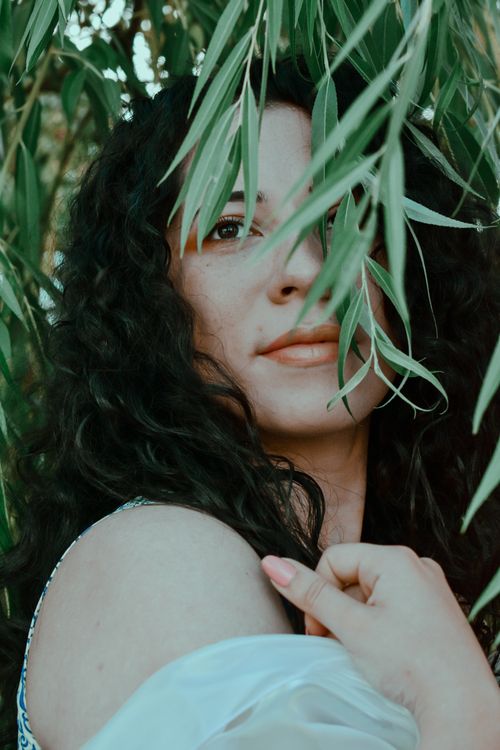 Free Graceful young female with dark wavy hair standing behind green plants in garden and looking away Stock Photo