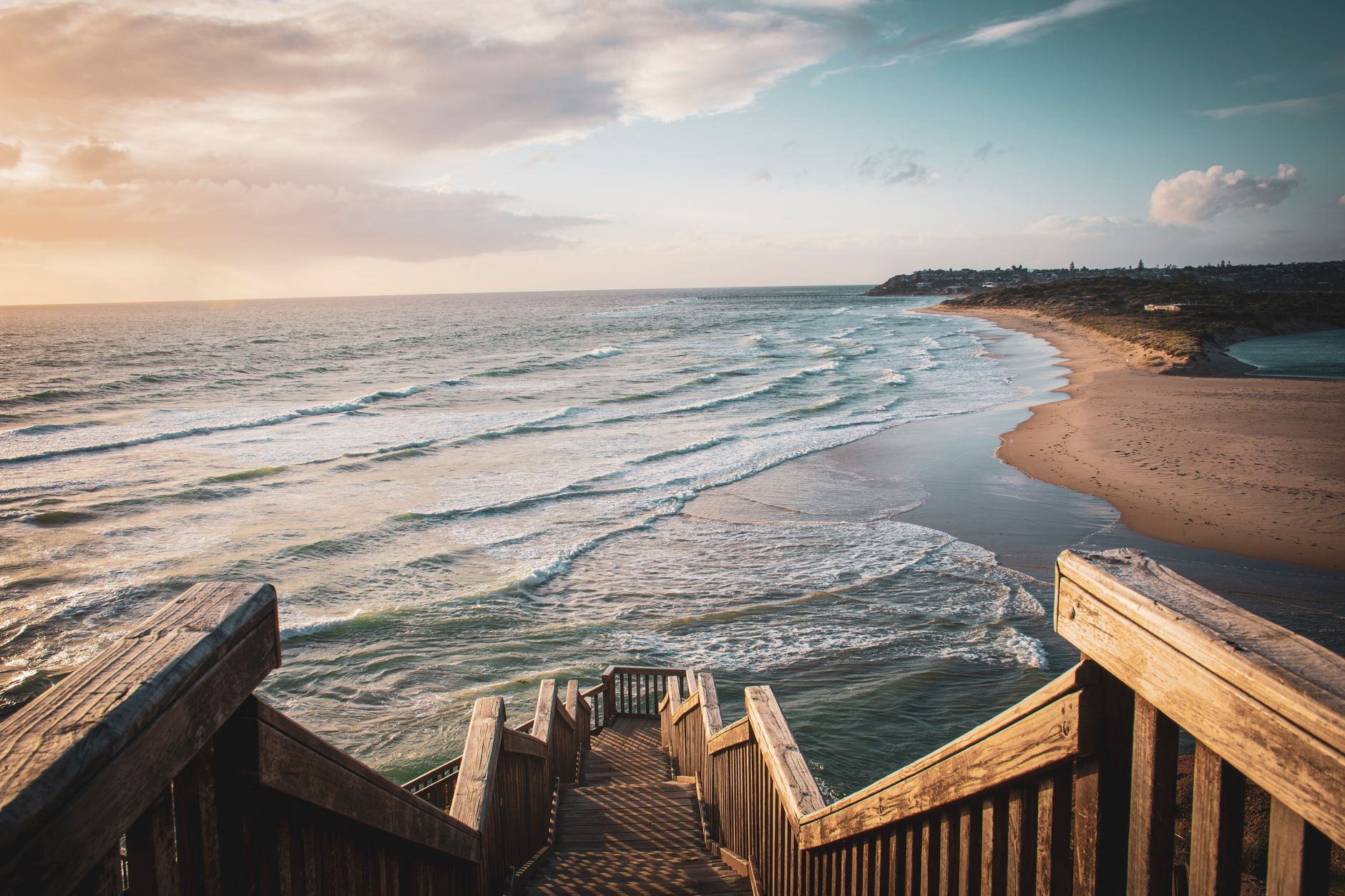 A picturesque view of the Adelaide beach with wooden stairs leading down, capturing waves and a sunset sky.