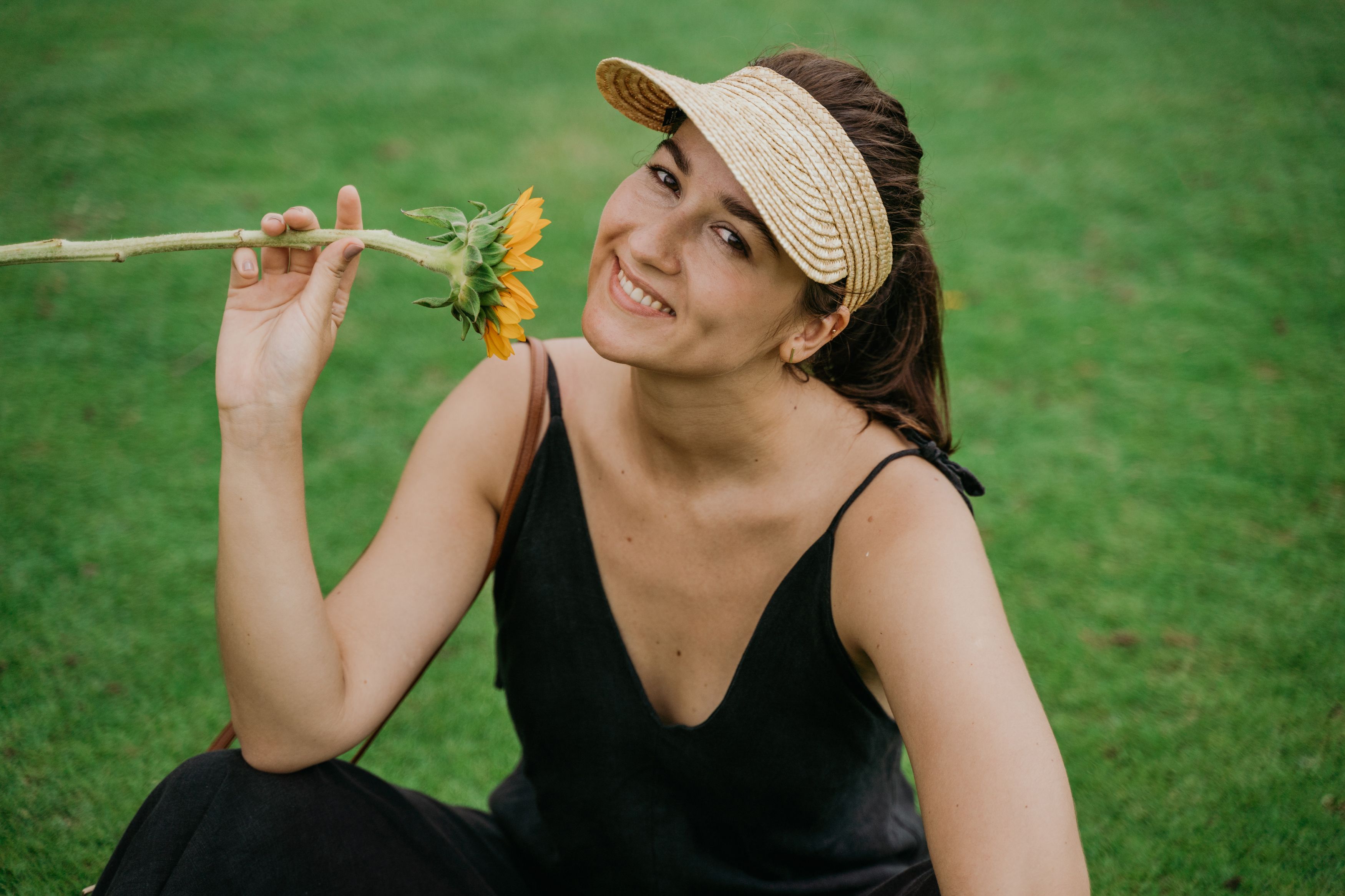 Smiling woman in a summer dress enjoys a sunflower while sitting on lush green grass.