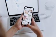 手拿智能手机展示画廊，背景是笔记本电脑，旁边放着一杯水