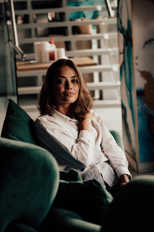 Beautiful positive female in white blouse resting on comfortable couch and touching shoulder while looking at camera contentedly