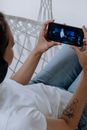 一名女子穿着蓝色牛仔裤、戴着耳机，在室内一边放松一边用智能手机看视频