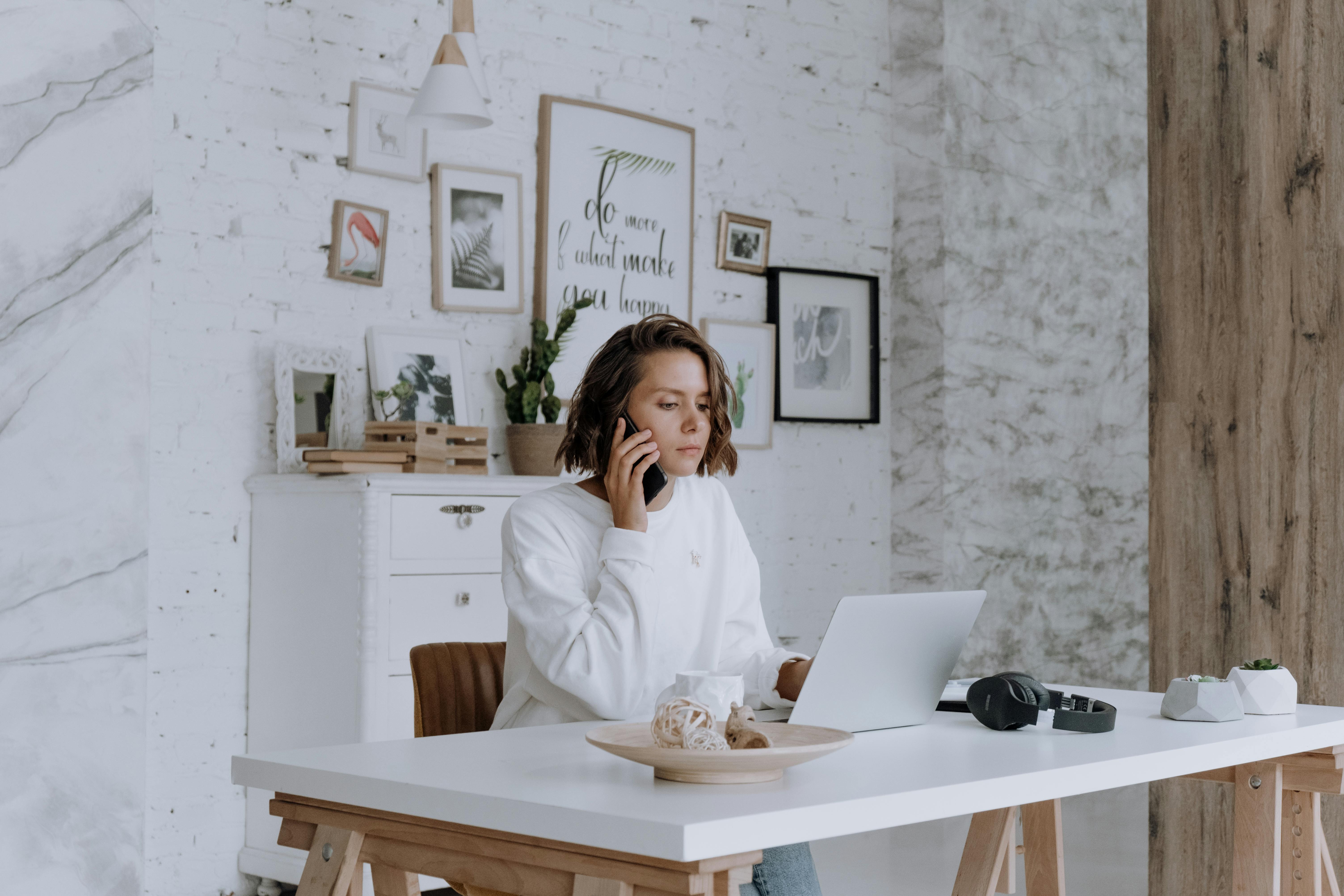 Woman talking on phone while using laptop in bright, modern home office with decor.