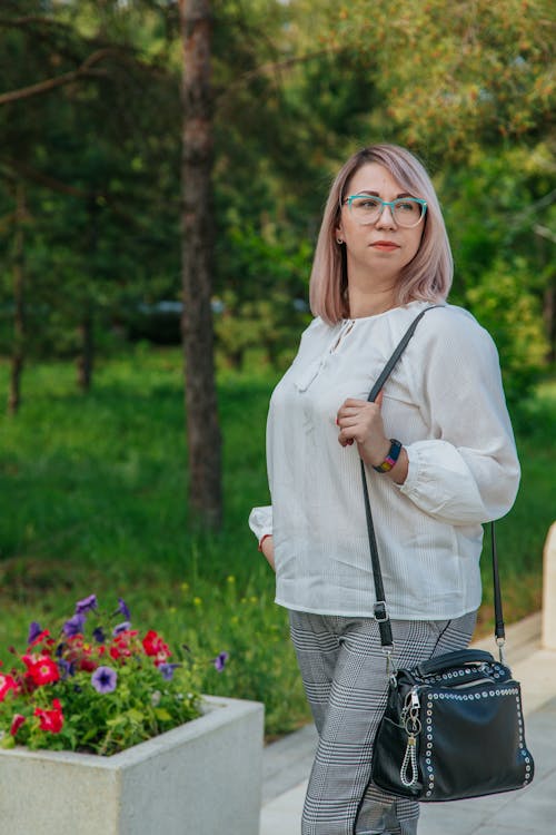 Young contemplative trendy female in eyewear with leather shoulder bag looking away near flowers and grass meadow with trees