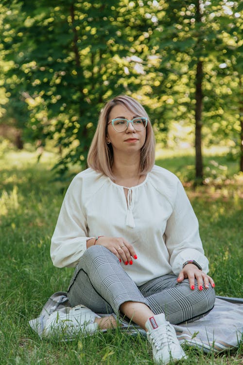 Young dreamy female in trendy clothes and eyeglasses sitting with crossed legs on fabric on grass meadow while looking away