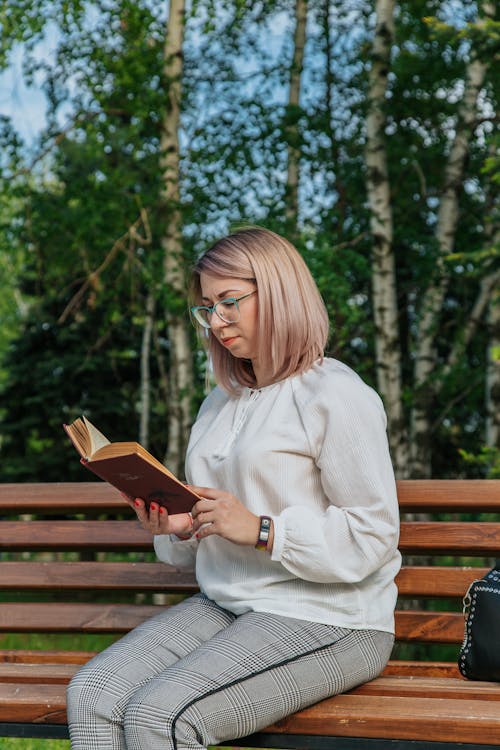 Young focused female in trendy wear reading textbook on wooden bench near trees in town