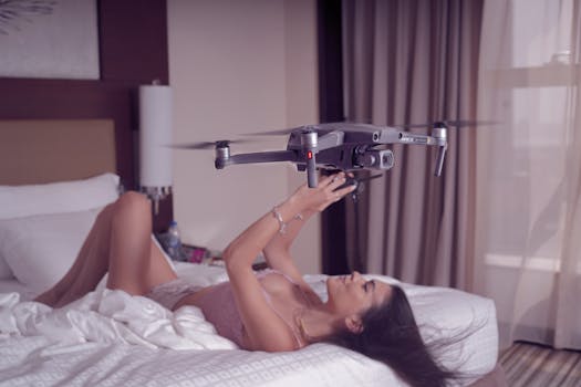 An adult woman lying on her bed indoors with a drone in her hands, surrounded by soft lighting.