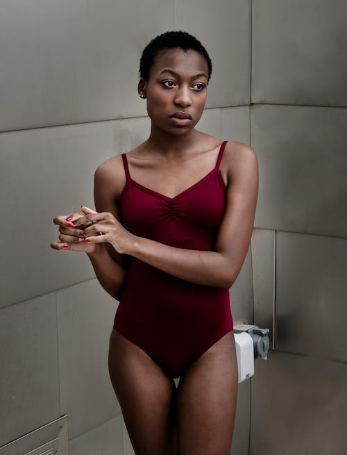 Thoughtful young slender African American lady with short hair in stylish bodysuit standing near gray wall and looking away