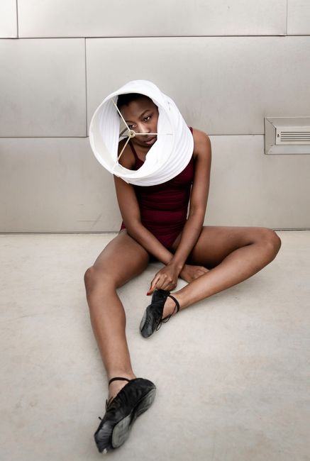 From above of exhausted young black female dancer in body suit and ballet shoes recreating on floor with lampshade on head and looking away pensively