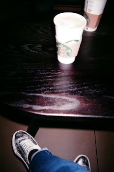 Two disposable coffee cups on a dark wooden table with a person wearing shiny sneakers visible.