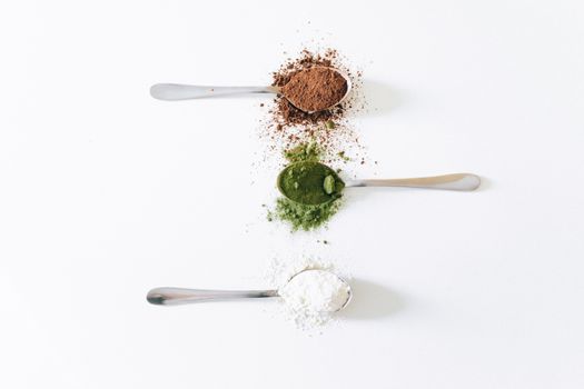 Top view of colorful assorted powders on spoons against white background.
