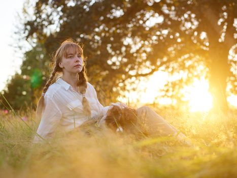 A woman sits in a sunlit field with her dog, enjoying nature's tranquility.