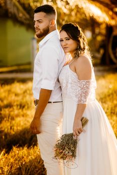 Side view of young ethnic bride in white wedding dress with bunch of flowers in hand embracing pensive groom while standing together on grassy meadow