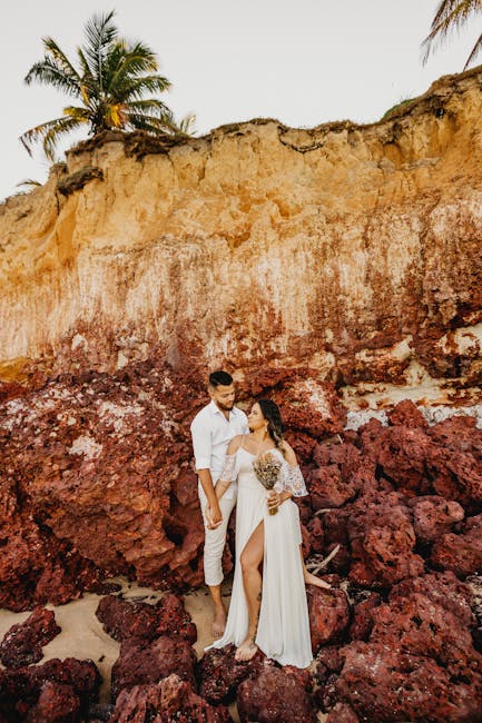 Full length of barefooted young female and male in white clothes holding hands while standing on rough rock near cliff during summer holidays in tropical country