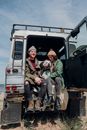 Man in Green Jacket Sitting on White Van With 2 Dogs
