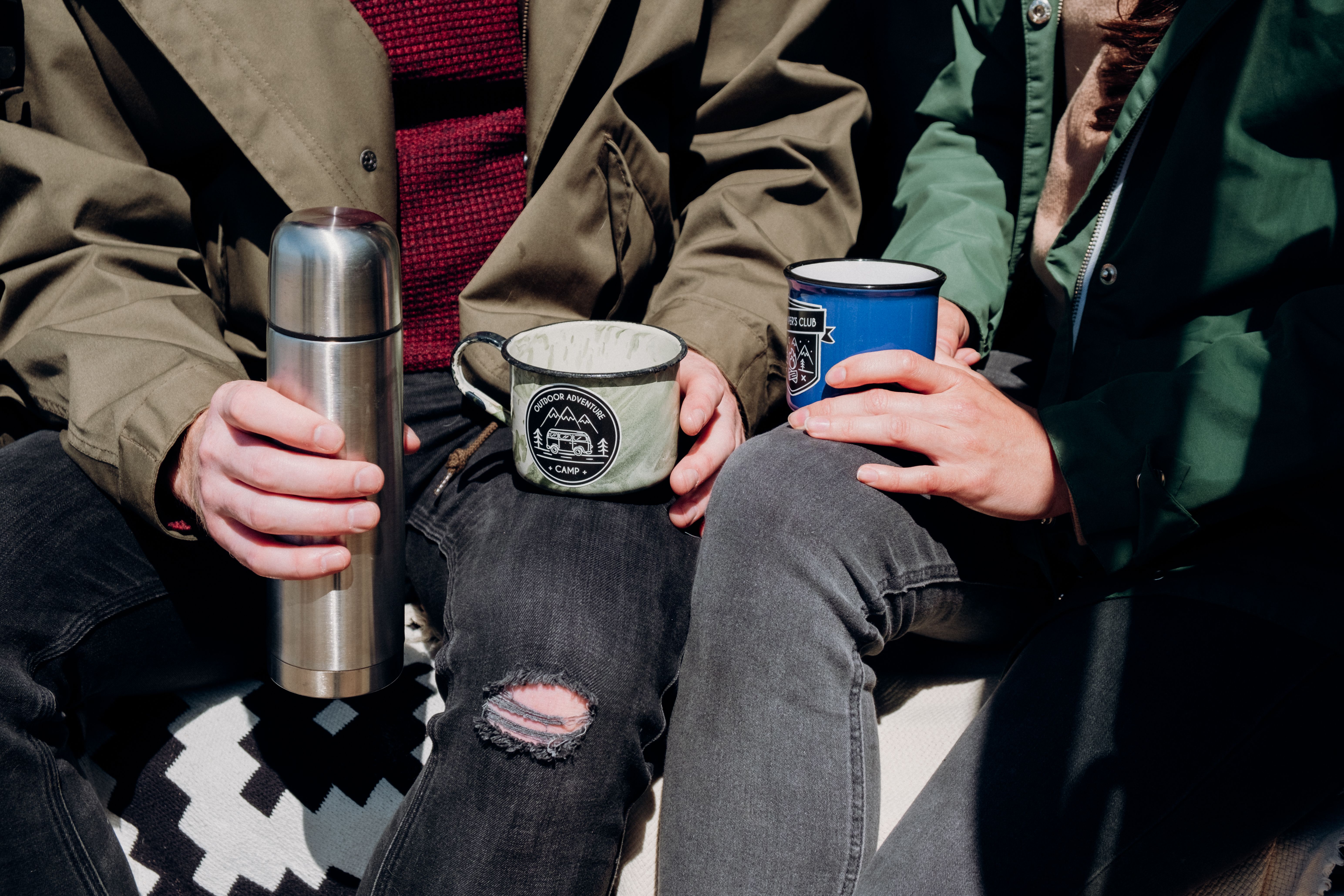 Casual outdoor moment with a couple holding mugs and a thermos, wearing casual jackets and denim jeans.