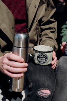 Close-up of hands holding a thermos and camp mug, embracing outdoor vibes.