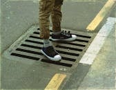 From above crop anonymous male legs in casual wear standing on asphalt road sewerage steel grating in daylight