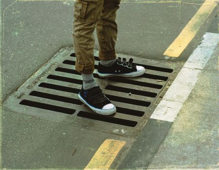 From above crop anonymous male legs in casual wear standing on asphalt road sewerage steel grating in daylight