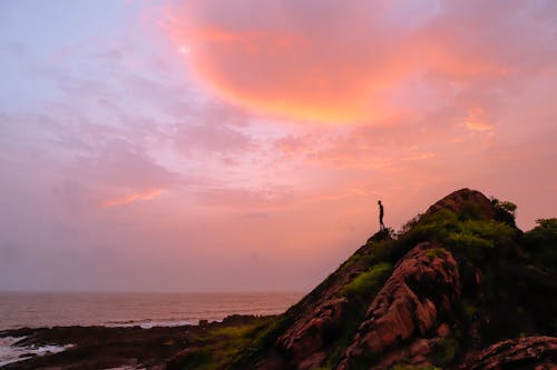 A Person Standing on Rock Formation Near Body of Water during Sunset
